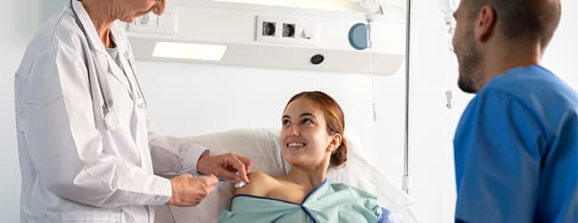 Médica atende paciente internada em leito hospitalar, acompanhada por outro profissional de saúde
